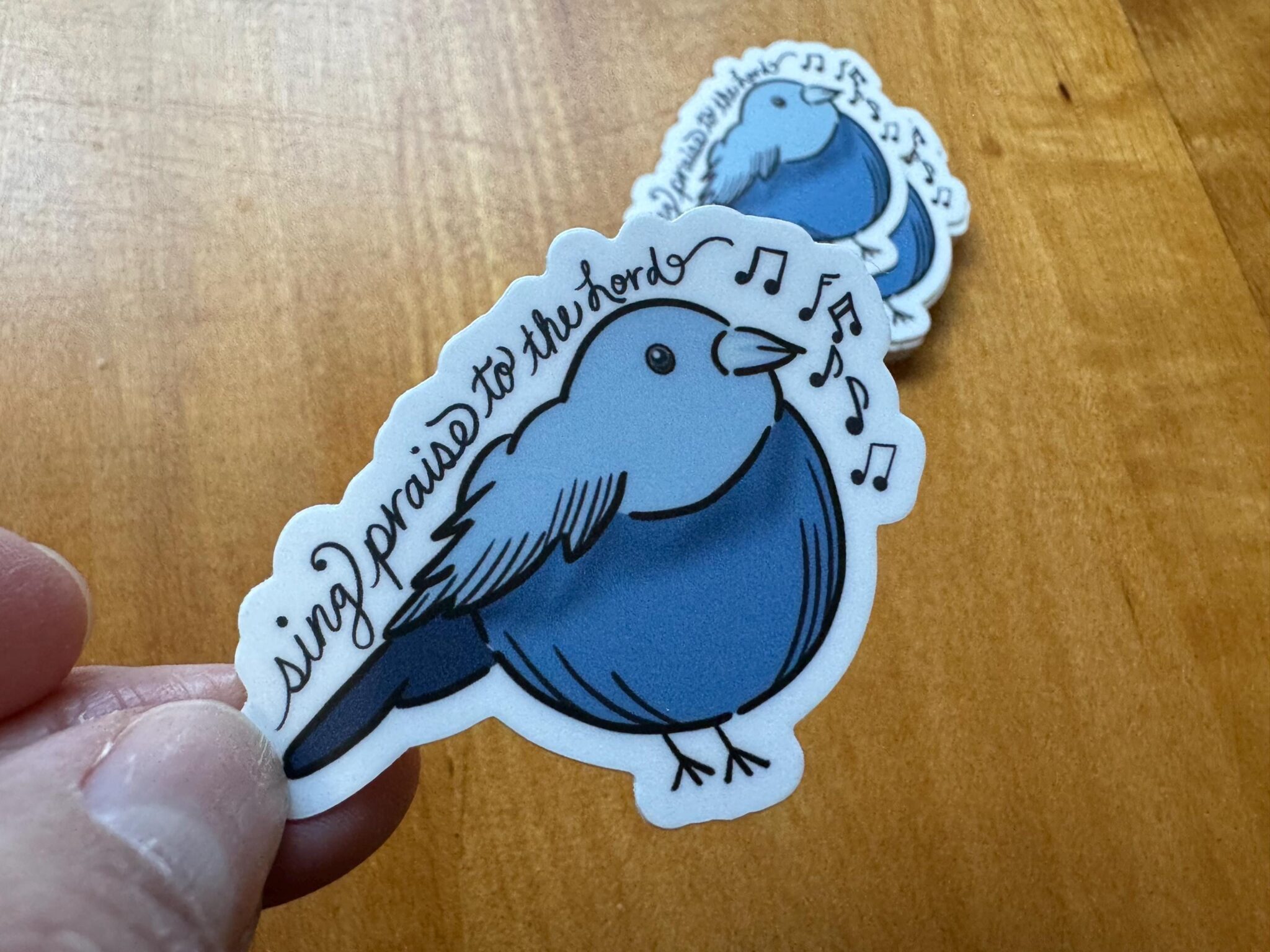 Blue bird sticker with musical notes and the phrase sing praise to the Lord held over wooden surface.
