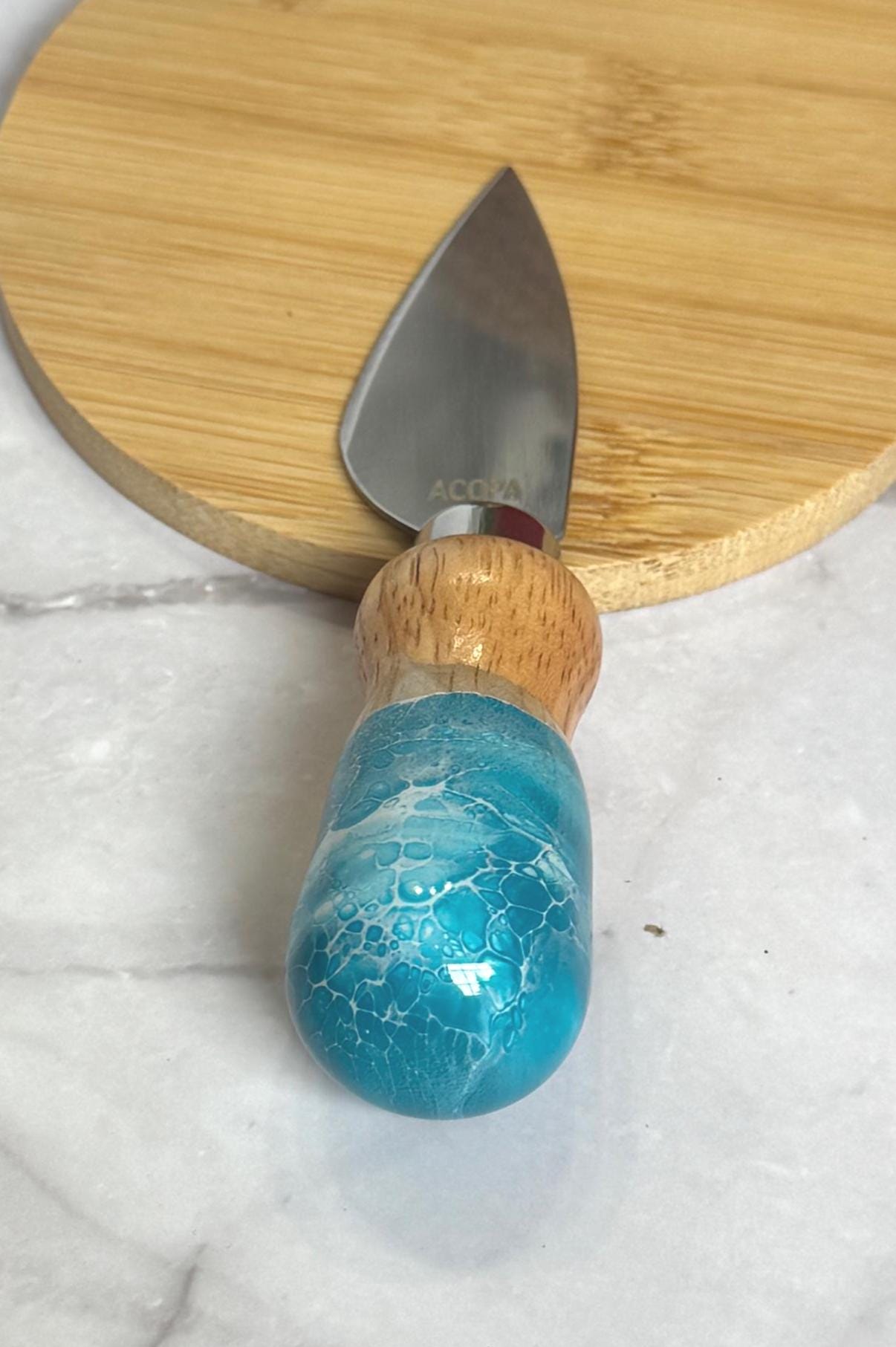 Cheese knife with blue resin and wooden handle on round wooden cutting board.