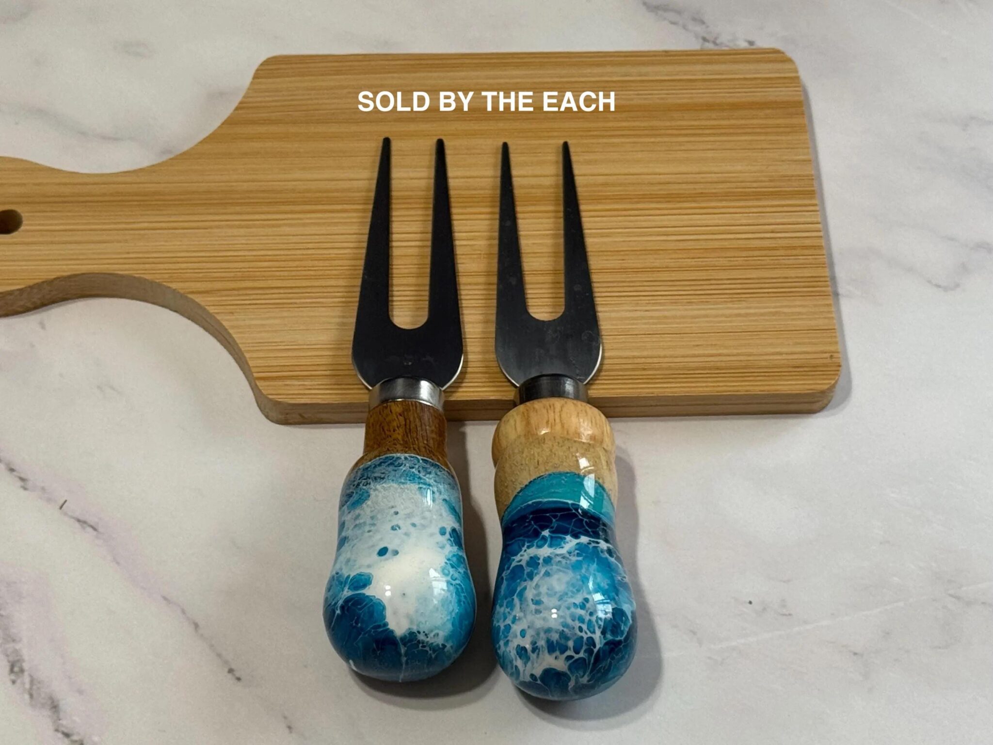Two cheese forks with blue and white marbled wooden handles on a bamboo cutting board.