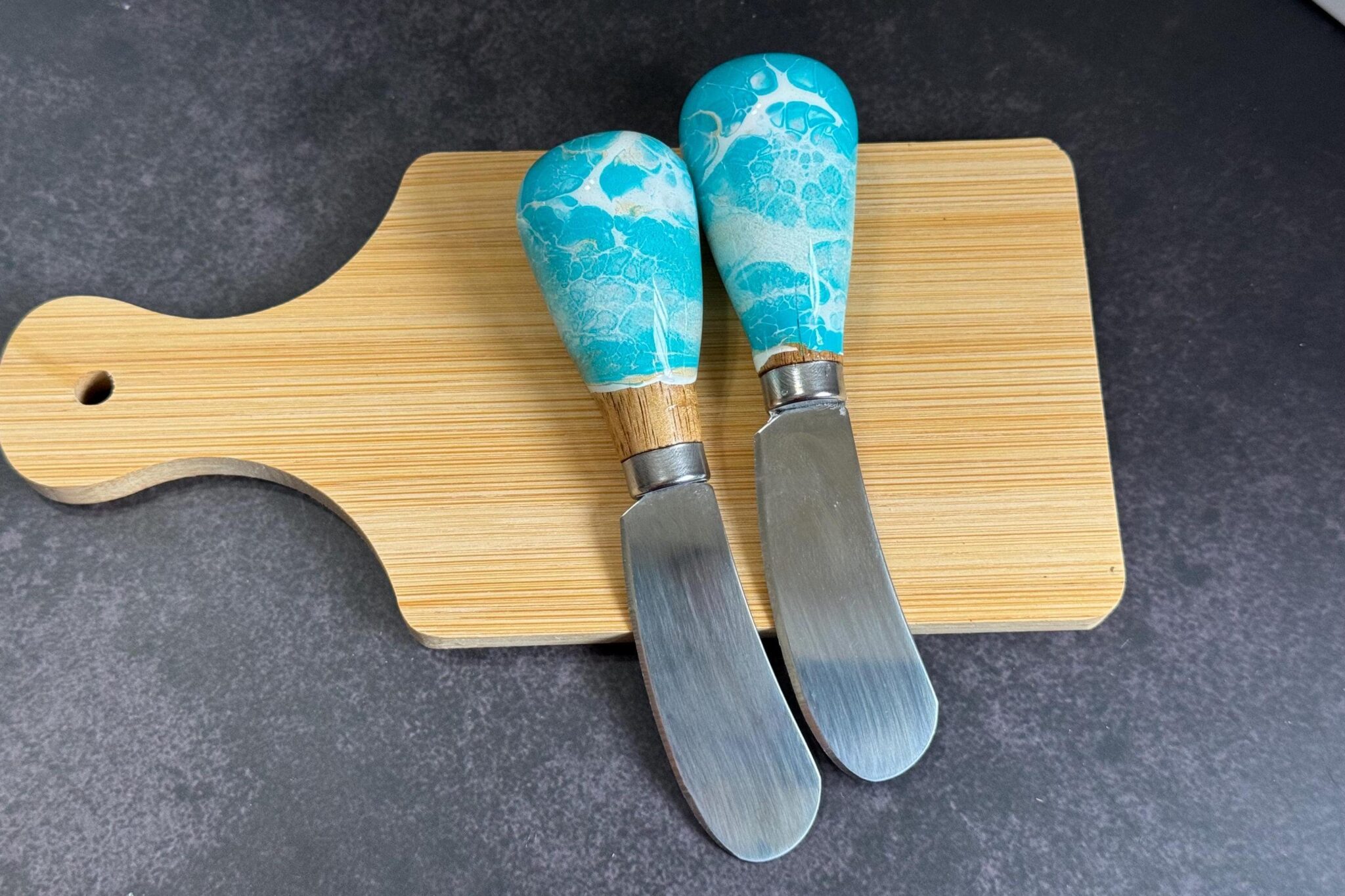Two cheese knives with blue and white handles on a wooden cutting board on a dark surface