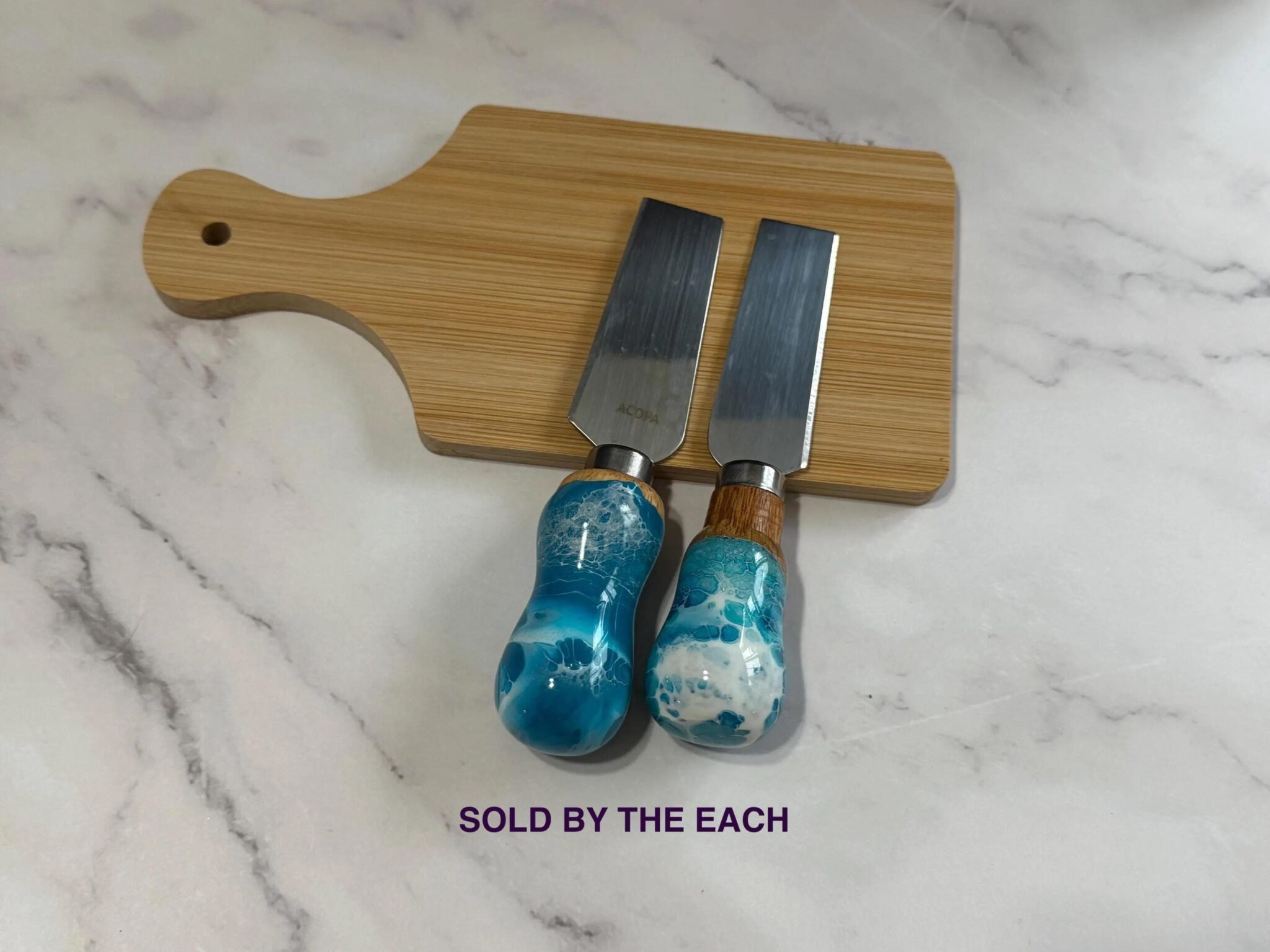 Cheese knives with blue and white resin handles on small wooden cutting board on marble surface with sold by each text.