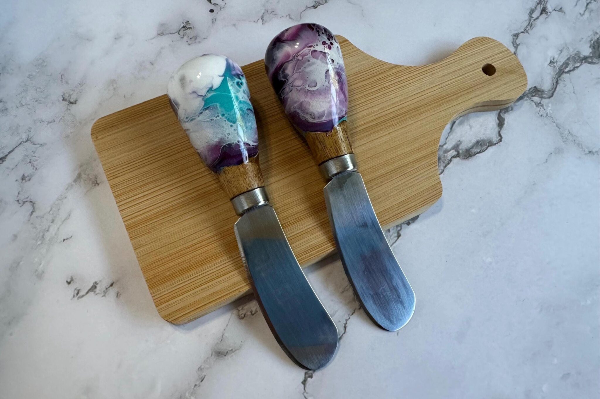 Two spreader knives with colorful resin and wood handles on a small wooden cutting board.