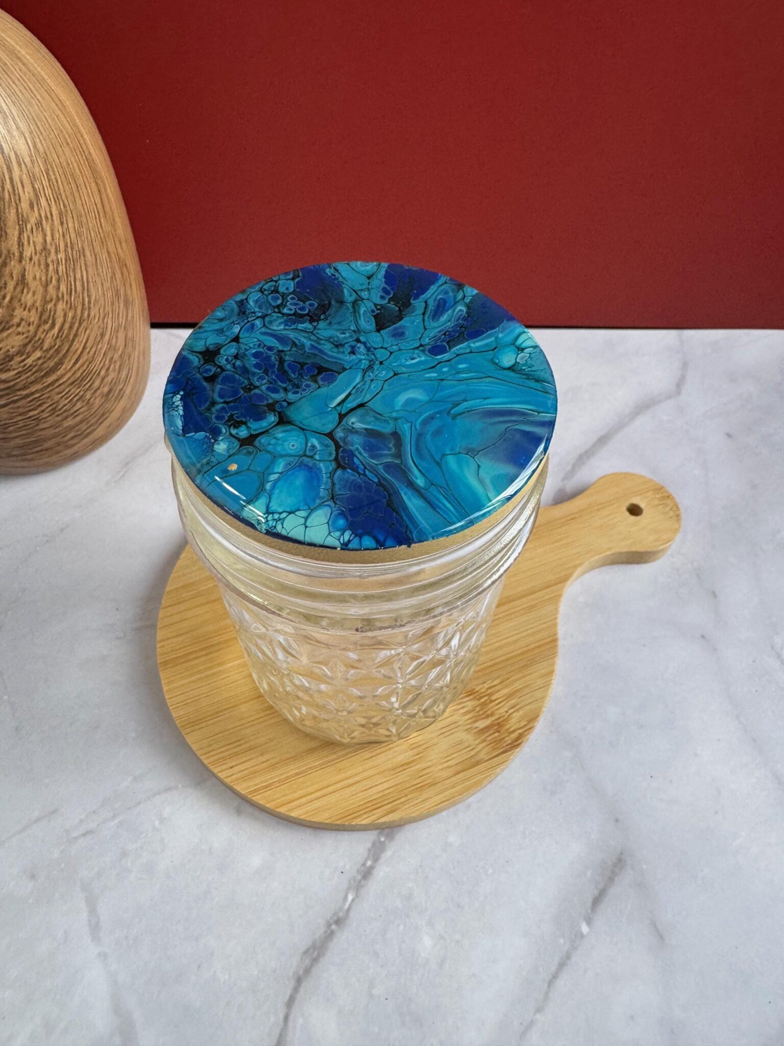 Glass jar with blue marbled lid on a small round wooden board on marble surface.