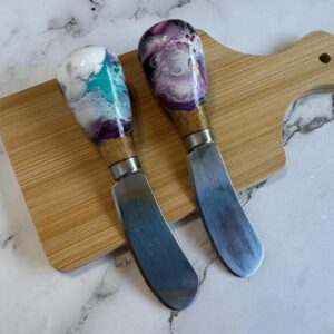 Two spreader knives with colorful resin and wood handles on a small wooden cutting board.