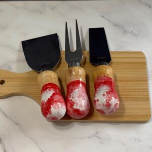 Three cheese knives with red and white handles on a wooden cutting board on a marble surface.