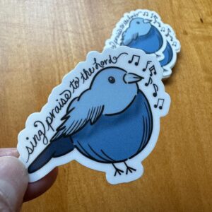 Blue bird sticker with musical notes and the phrase sing praise to the Lord held over wooden surface.
