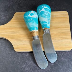 Two cheese knives with blue and white handles on a wooden cutting board on a dark surface