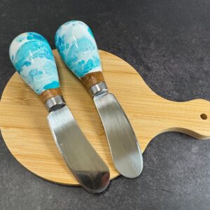 Two cheese knives with blue patterned handles on a wooden cutting board on a dark surface