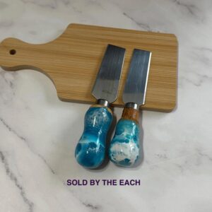 Cheese knives with blue and white resin handles on small wooden cutting board on marble surface with sold by each text.