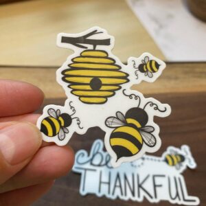 Hand holding a sticker of a yellow beehive and three bees with a thankful sticker in background