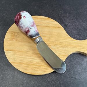 Round wooden cutting board with a cheese spreader featuring a marbled handle on dark surface