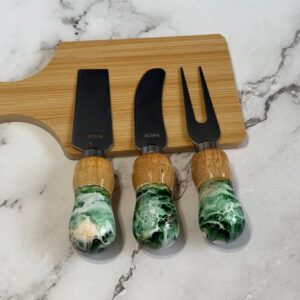 Three cheese knives with wooden and green marbled handles on a wooden cutting board on marble surface.