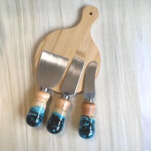 Wooden cutting board with three cheese knives featuring wooden and blue resin handles.