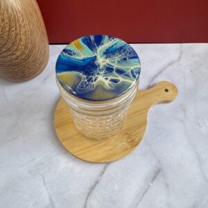 Clear glass jar with blue and gold lid on round wooden board on white marble surface.