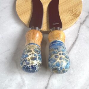 Two cheese knives with wooden and blue crackled handles on a round wooden cutting board.