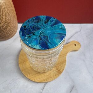 Glass jar with blue marbled lid on a small round wooden board on marble surface.