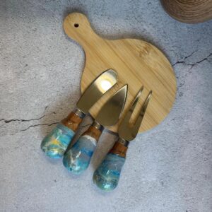 Wooden serving board with three cheese utensils with blue and wooden handles on gray surface