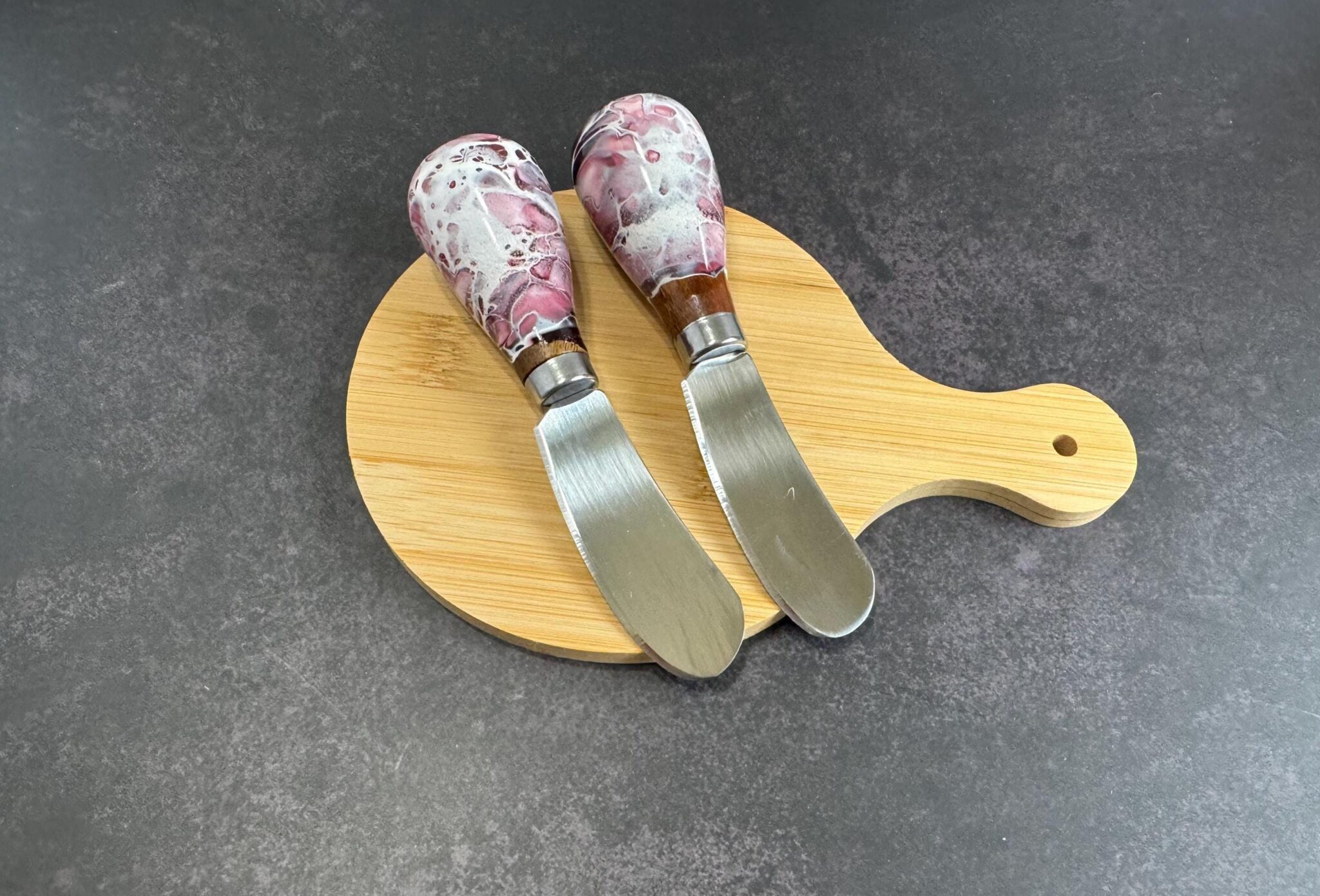 Two cheese spreader knives with marbled pink and white handles on a round wooden cutting board.
