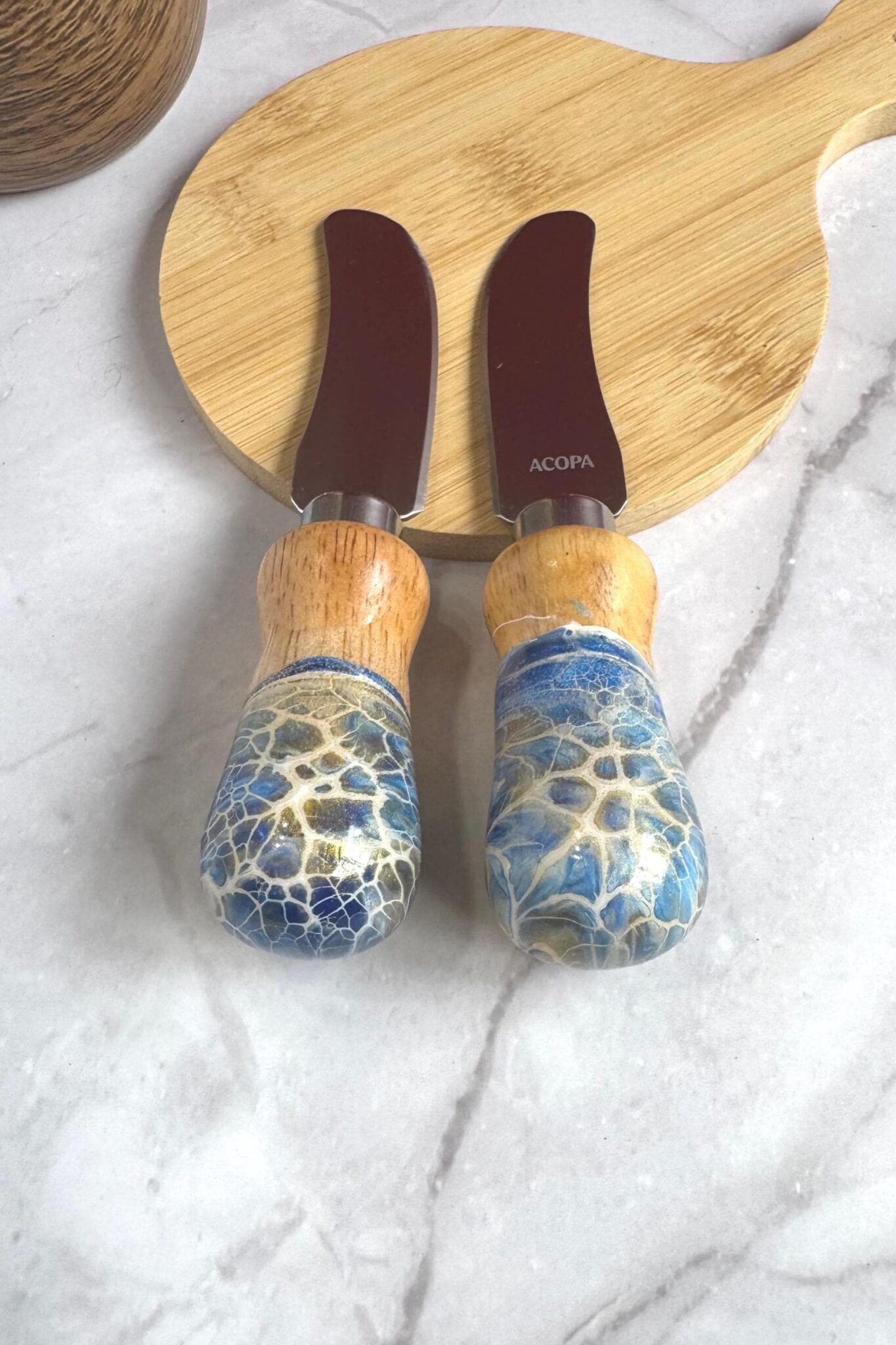 Two cheese knives with wooden and blue crackled handles on a round wooden cutting board.