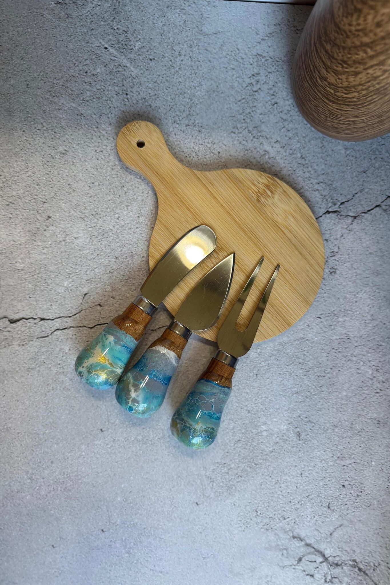Wooden serving board with three cheese utensils with blue and wooden handles on gray surface