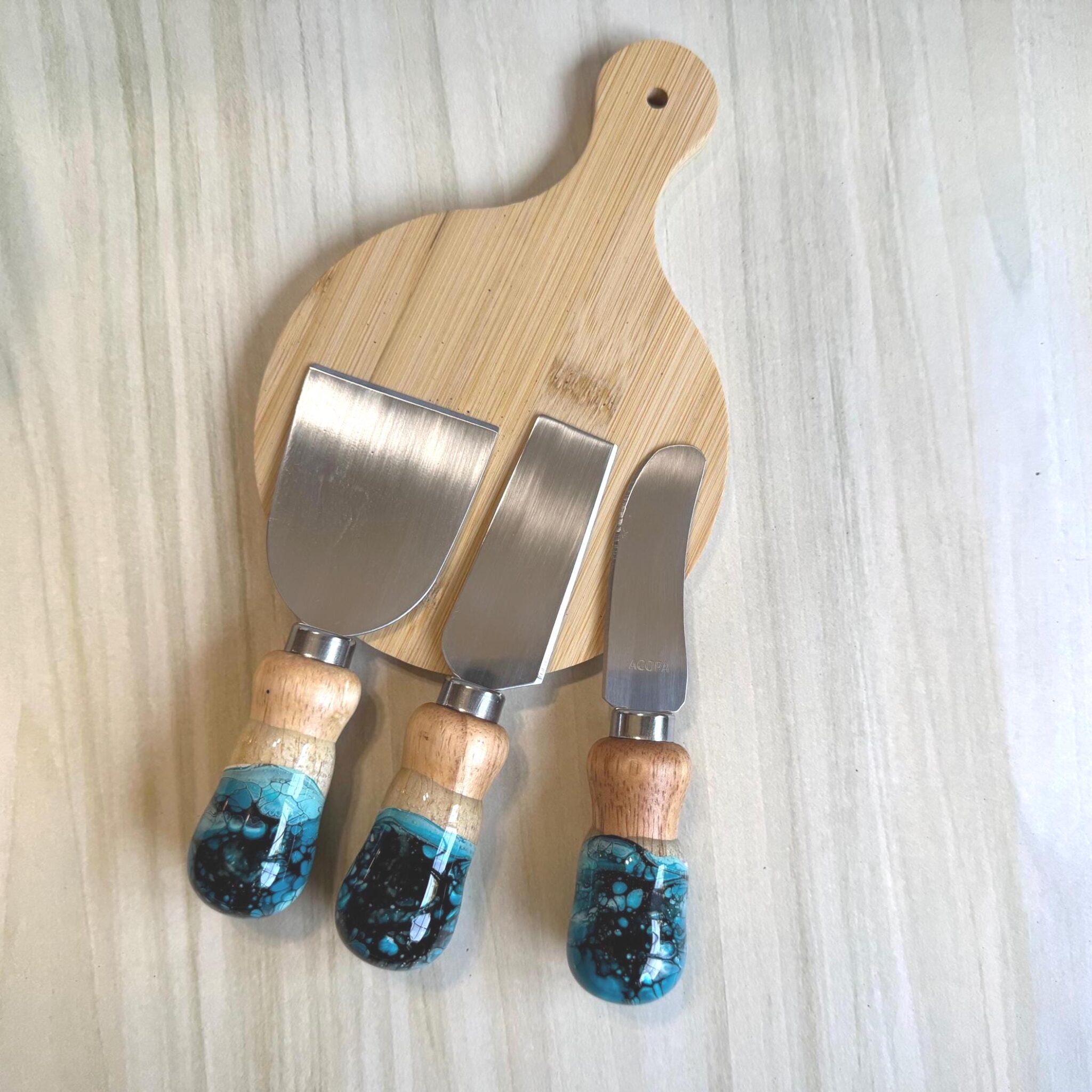 Wooden cutting board with three cheese knives featuring wooden and blue resin handles.